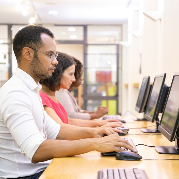 Diverse group of students taking online tests in computer class. Line of man and women in casual sitting at table, using desktops, typing, looking at monitor. Training course concept